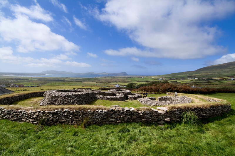 A historic stone ring fort (caiseal or stone cashel) situated on a grassy hill overlooking the green fields of the Dingle Peninsula and a coastal bay under a blue sky with white clouds.
