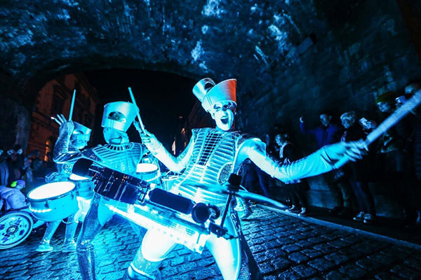 Three performers in metallic silver costumes glowing with bright blue LED lights play drums under a dark stone archway at night.
