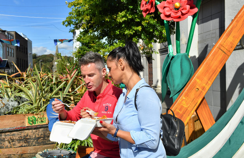 A man and woman sit outdoors on a sunny day, enjoying street food from white takeaway containers. The man wears a red quarter-zip sweater and the woman wears a light blue shirt with a black backpack. They are surrounded by vibrant green plants and large decorative red flower sculptures.