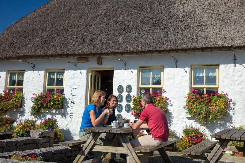 Three people sitting at a wooden picnic table enjoying tea outside Teach nan Phaidi, a traditional white-washed thatched cottage cafe on Inishmore, Aran Islands.
