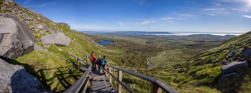 High-angle panoramic view from a wooden staircase on Cuilcagh Mountain, looking down at a long boardwalk winding through a green valley toward a distant lake under a clear blue sky.