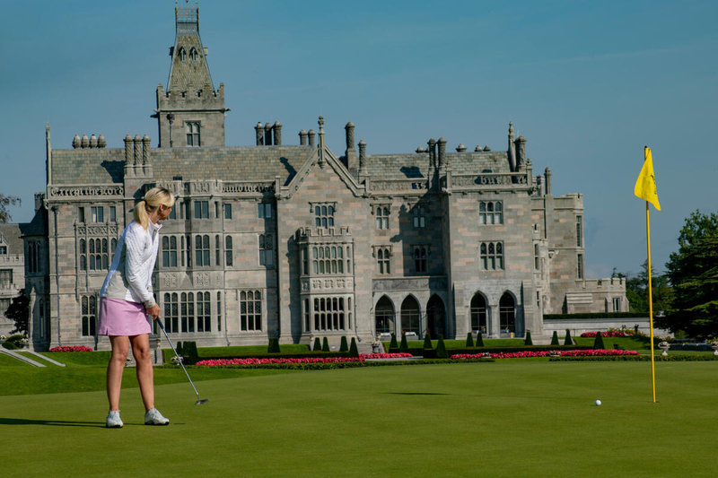 A female golfer in a pink skirt and white jacket putts a golf ball on a lush green toward a yellow flag, with the historic, turreted Adare Manor stone building in the background under a clear blue sky.