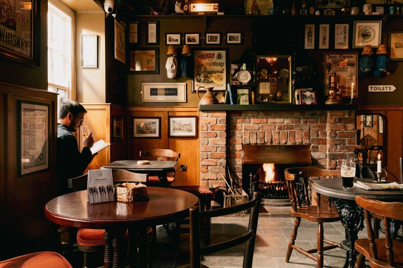 The interior of a traditional, dimly lit bar at the Harbourview Hotel in Carnlough. A man sits by a window on the left, reading a book and tasting whiskey. In the center, a warm fire glows in a brick fireplace, surrounded by wood-paneled walls covered in vintage whiskey advertisements, framed photos, and memorabilia. Dark wooden tables and chairs fill the room, with a pint of stout and a lit candle on a table in the foreground, creating a quiet and inviting atmosphere.