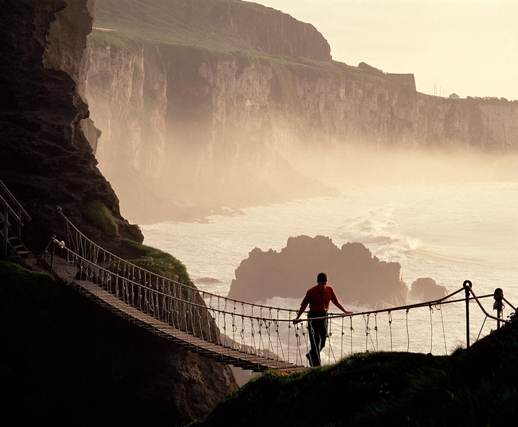 A dramatic, silhouetted view of the Carrick-a-Rede Rope Bridge in County Antrim at sunset. A lone man walks across the narrow wooden plank bridge, which spans a deep chasm between high, rugged cliffs. In the background, the ocean waves crash against rocky outcrops, and a soft, golden mist rises against the towering limestone cliffs, creating a hazy and ethereal coastal scene.