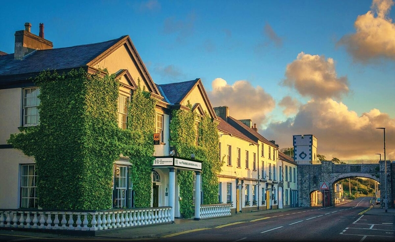 A side-profile view of the Harbourview Hotel in Carnlough, Northern Ireland, featuring a grand white building partially covered in lush green ivy. The scene includes a white balustrade in the foreground, a quiet street leading to a historic stone arch bridge in the background, and a warm sunset glow across the clouds.