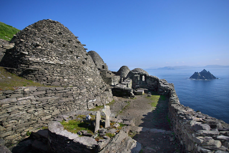A group of ancient dry-stone beehive huts (clocháns) on a cliffside terrace of Skellig Michael, overlooking the blue Atlantic Ocean with Little Skellig in the distance.